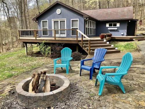 A cozy cabin with a deck, surrounded by trees, features a fire pit and colorful chairs in the foreground.