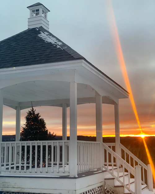 A white gazebo with a Christmas tree inside, overlooking a sunset with vibrant orange and yellow hues.