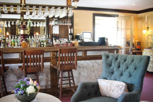 Cozy bar area with a marble counter, wooden stools, and a comfortable green armchair beside a small table with flowers.