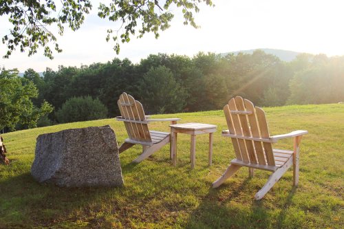 Two wooden Adirondack chairs and a small table sit on a grassy hill, surrounded by trees and bathed in warm sunlight.