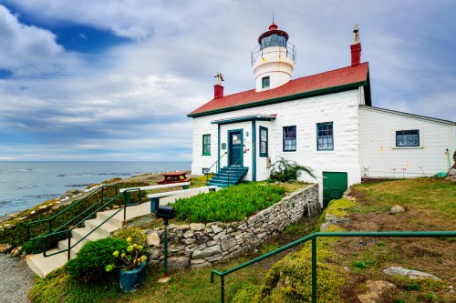 A charming white lighthouse with a red roof, surrounded by greenery, overlooking the ocean under a cloudy sky.