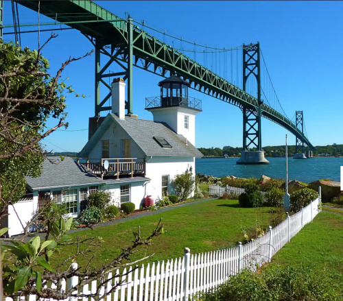 A white lighthouse and house with a green bridge in the background, overlooking a calm body of water under a clear blue sky.