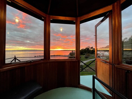 A cozy wooden room with large windows showcasing a vibrant sunset over the water and a scenic landscape outside.