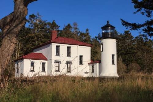 A white lighthouse with a red roof, surrounded by greenery and trees under a clear blue sky.