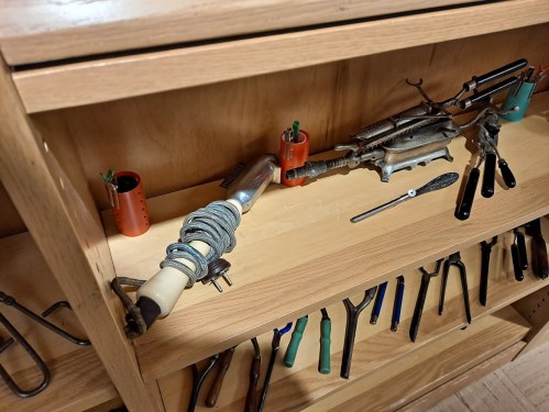 A wooden shelf displaying various tools, including a wrapped handle tool and several pliers and clamps.