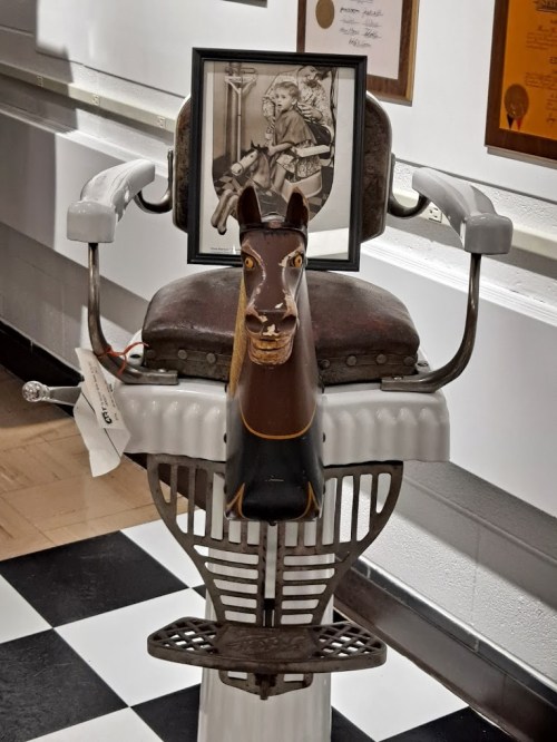 A vintage barber chair with a horse-shaped headrest and a framed photo of a child with a horse in the background.