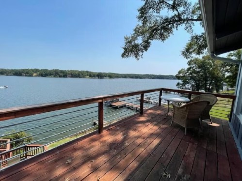 A wooden deck overlooking a calm lake, with a table and chair, surrounded by trees under a clear blue sky.