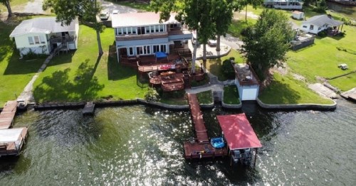 Aerial view of a lakeside property with a house, dock, and green lawn, surrounded by water and trees.