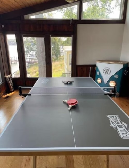 A ping pong table in a bright room with large windows, overlooking a lake, and a vintage blue cabinet nearby.