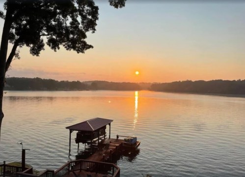 Sunset over a calm lake, with a dock and boat, surrounded by trees and misty hills in the background.
