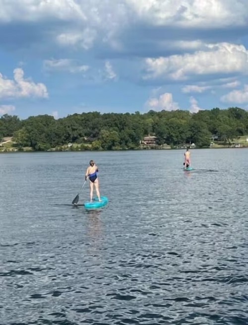 Two people paddleboarding on a calm lake under a blue sky with fluffy clouds.