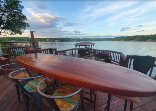 A wooden table with metal chairs overlooks a serene lake and a cloudy sky from a deck.