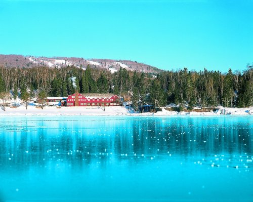 A red building by a frozen lake, surrounded by snow-covered trees and mountains under a clear blue sky.