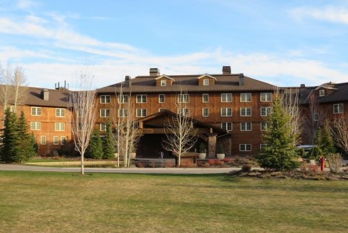 A large, rustic brick building with a covered entrance, surrounded by trees and a grassy area under a blue sky.