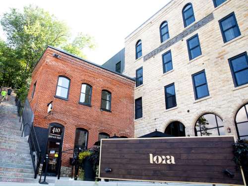 Historic brick and stone buildings with a modern sign for "Lora" in a vibrant urban setting. Steps lead up beside them.