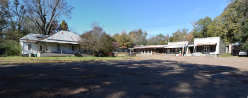 A quiet, rural street scene featuring an old house and a row of abandoned storefronts under a clear blue sky.