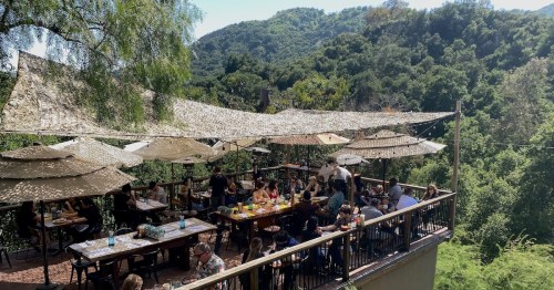A lively outdoor restaurant with shaded tables, surrounded by lush greenery and mountains in the background.