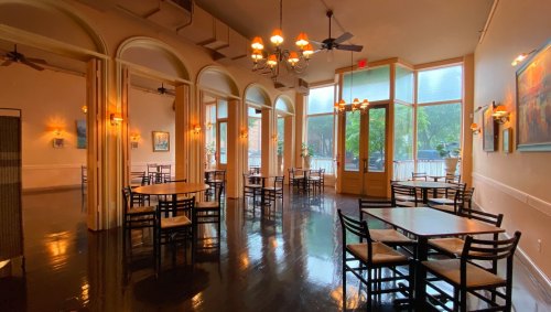 A spacious, softly lit restaurant with wooden tables, chairs, and large windows overlooking greenery outside.