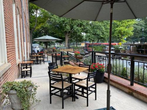 Outdoor patio with tables and chairs, surrounded by greenery and flowers, under large umbrellas. Brick building in background.