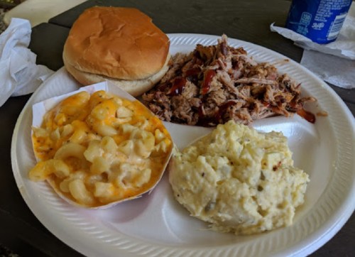 A plate with pulled pork, macaroni and cheese, mashed potatoes, and a bun, served outdoors.