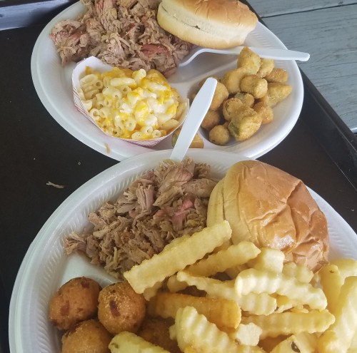 Two plates of food featuring pulled pork, macaroni and cheese, hushpuppies, crinkle-cut fries, and sandwich buns.