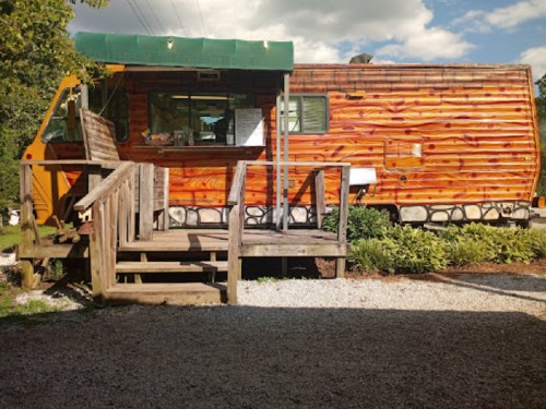 A colorful wooden food truck with a green awning, set on a gravel path surrounded by greenery.