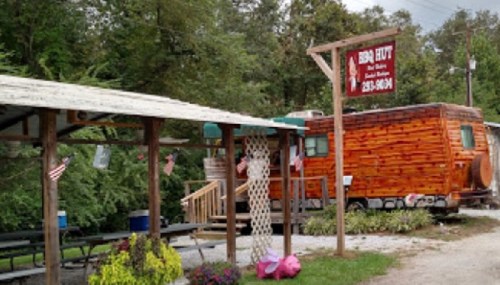 A rustic BBQ hut with a wooden trailer, surrounded by trees and picnic tables, featuring a sign and decorative flags.
