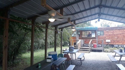A covered outdoor area with picnic tables, a fan, and a food truck in the background surrounded by greenery.