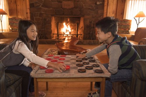 Two children play checkers by a cozy fireplace in a wooden cabin, smiling and focused on the game.