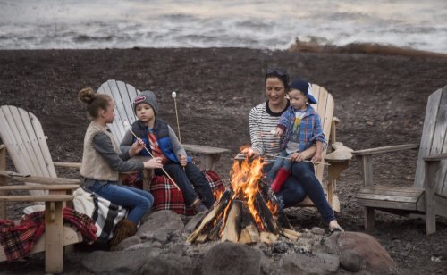 A woman and three children roast marshmallows by a campfire, seated on wooden chairs near a beach.