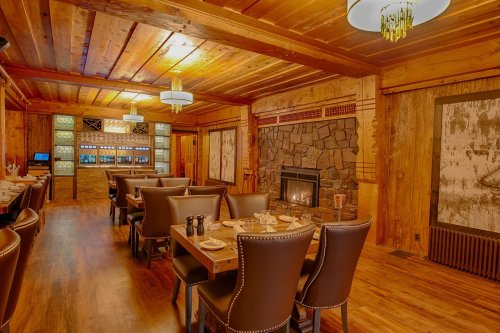 Cozy wooden restaurant interior with tables, leather chairs, a stone fireplace, and warm lighting.