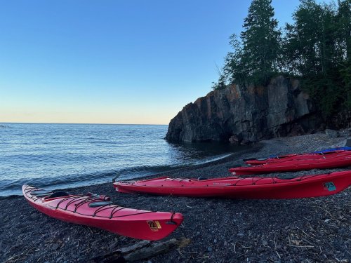 Red kayaks lined up on a rocky beach beside calm water, with a cliff and trees in the background under a clear blue sky.