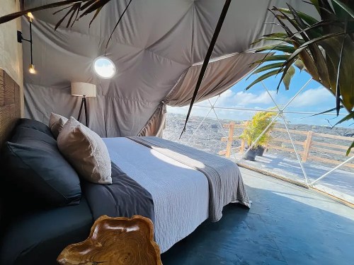Cozy interior of a geodesic dome with a bed, natural light, and a view of the outdoors through large windows.