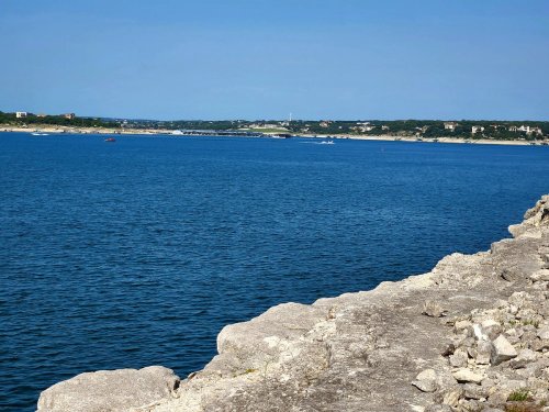 A serene view of a blue lake with rocky shoreline and distant hills under a clear blue sky.