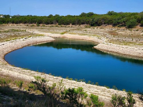 A serene, blue pond surrounded by dry, rocky terrain and sparse greenery under a clear blue sky.