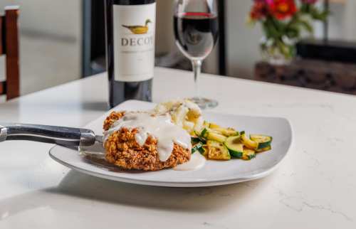 A plate of fried chicken with creamy sauce, served with zucchini and a glass of red wine in the background.