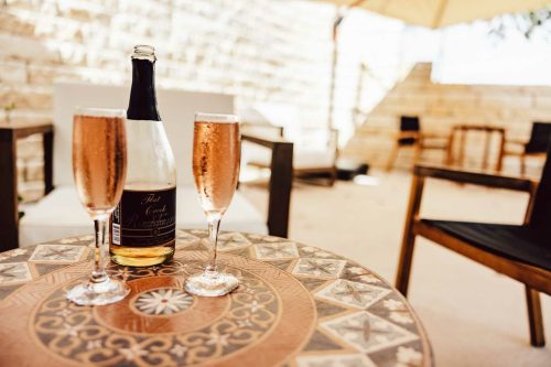 Two glasses of sparkling rosé sit on a decorative table beside a bottle, with a sunny outdoor setting in the background.