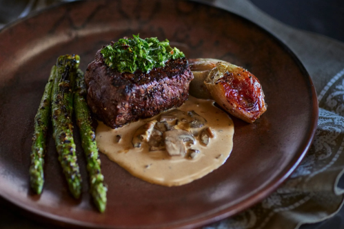 A plate featuring a grilled steak topped with herbs, asparagus, roasted tomato, and a creamy mushroom sauce.