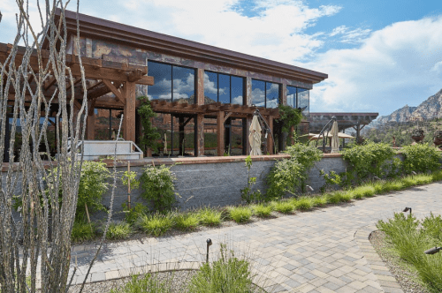 A modern building with large windows, surrounded by greenery and a stone pathway, set against a mountainous backdrop.