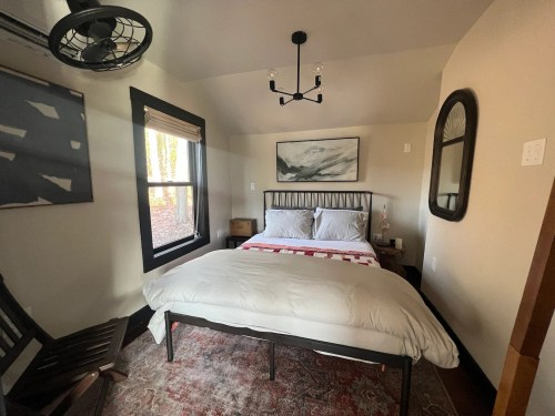 Cozy bedroom with a bed, wooden chair, and large windows letting in natural light. Decor includes a painting and mirror.