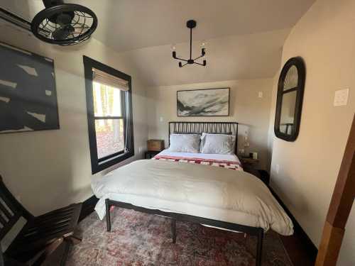 Cozy bedroom with a bed, wooden chair, and large windows letting in natural light. Decor includes a painting and mirror.