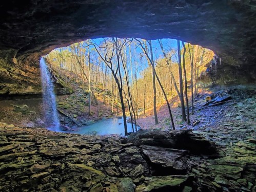 A scenic view from a cave opening, featuring a waterfall, trees, and a tranquil pool of water below.