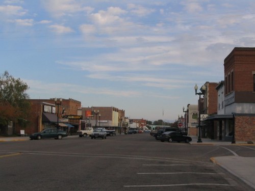 A quiet small-town street with brick buildings, parked cars, and a cloudy sky in the background.
