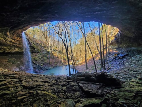 A scenic view from inside a cave, featuring a waterfall, trees, and a tranquil pool of water.