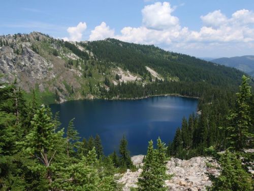A serene mountain lake surrounded by lush green trees and rocky hills under a blue sky with fluffy clouds.