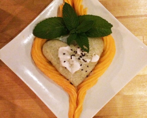 A heart-shaped dish with creamy topping, surrounded by vibrant orange sauce and fresh mint leaves on a white plate.