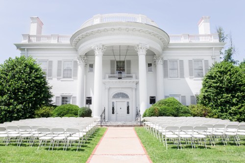A grand white mansion with columns, flanked by green bushes, and rows of empty chairs set up for an outdoor event.