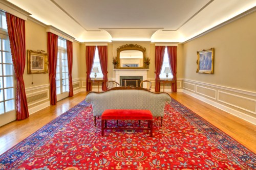 Elegant room with a red rug, vintage furniture, large windows, and decorative curtains, featuring a fireplace and artwork.