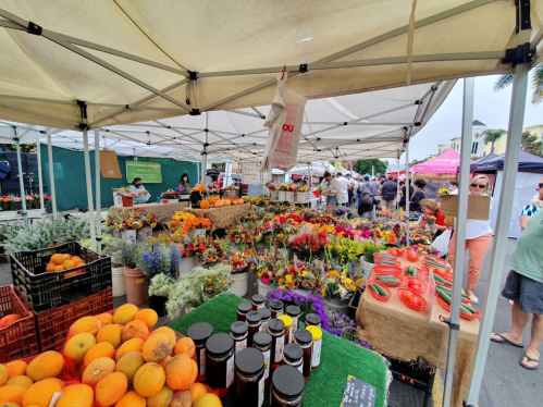 A vibrant farmers market with colorful flowers, fruits, and jars of preserves under tents, bustling with shoppers.