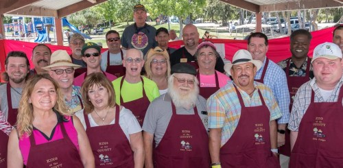 A diverse group of people in maroon aprons smiling together under a pavilion at a community event.